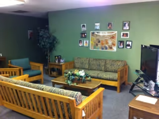 A cozy living room with wooden framed sofas and chairs featuring patterned cushions. A wooden coffee table with a floral centerpiece is in the center. The walls are painted green and decorated with framed pictures and a large floral artwork. A large flat-screen TV is positioned on a stand to the right side of the room.