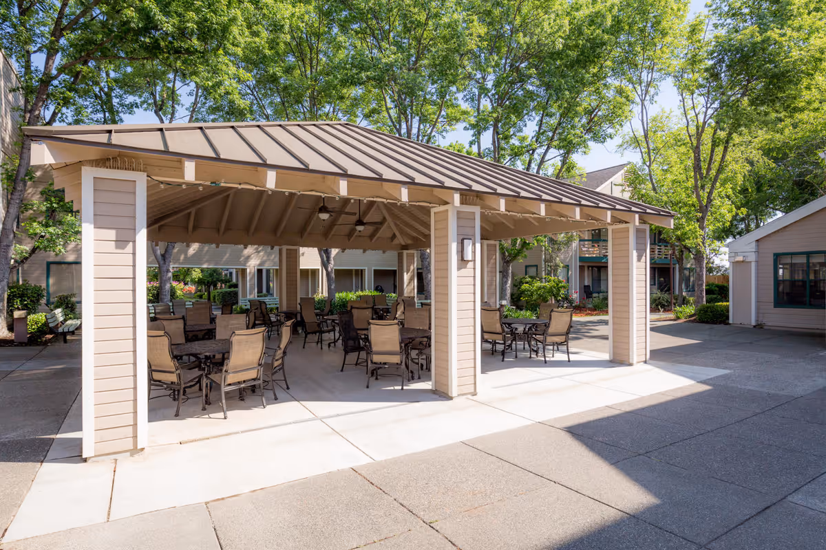 Covered outdoor pavilion with tables and chairs in a shaded courtyard surrounded by trees and buildings.