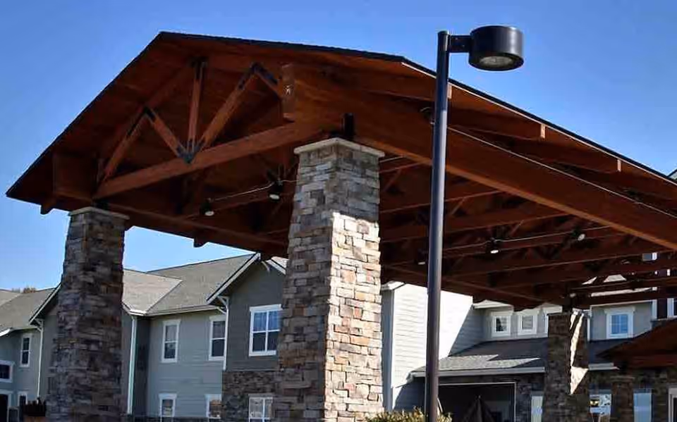 Outdoor covered pavilion structure with wooden beams and stone pillars, located in front of a residential building under a clear blue sky.