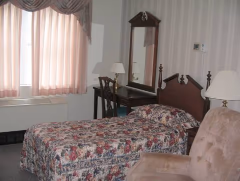 A floral-patterned bed with headboard, dresser mirror, desk, lamps, and pink-curtained window in a bedroom.