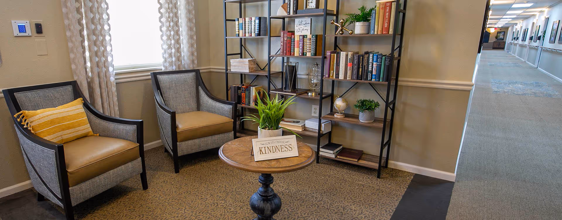 A small lounge area with two armchairs, a round table holding a potted plant and a 'Kindness' sign in front of a bookshelf and an adjacent hallway.