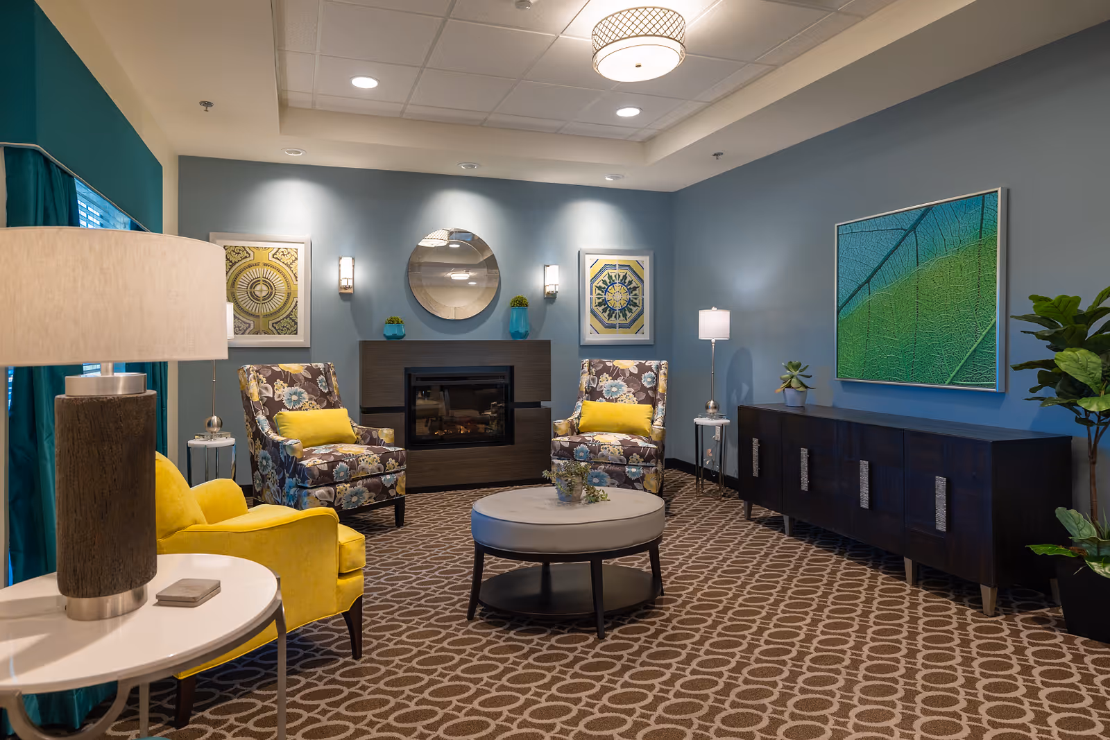 A cozy living room with two floral patterned armchairs with yellow cushions, a yellow armchair, a round ottoman coffee table, a modern fireplace with a round mirror above it, framed artwork on the walls, a dark wooden sideboard, and various lamps and plants. The walls are painted blue and the carpet has a geometric pattern.