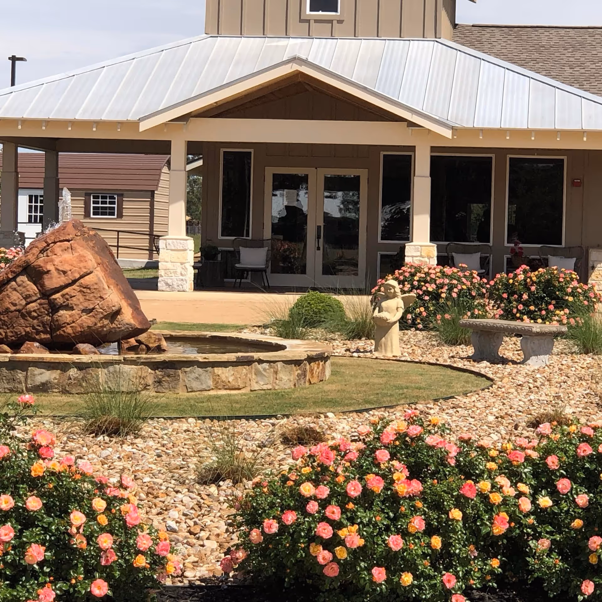 Covered entrance of a senior living building with a rock fountain, statue, stone bench, and colorful rose bushes in the foreground.