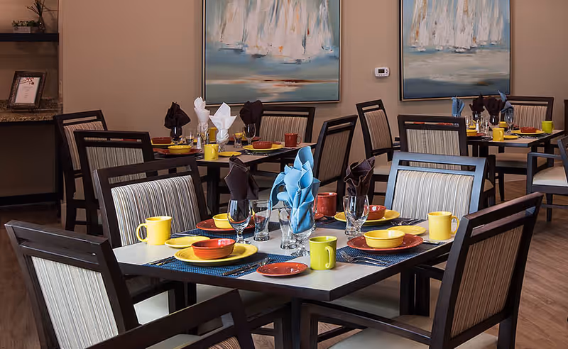A dining room with multiple tables set for a meal. Each table has colorful plates, bowls, mugs, and folded napkins in various colors including yellow, red, blue, and brown. The chairs have striped upholstery, and there are two abstract paintings on the beige walls in the background.