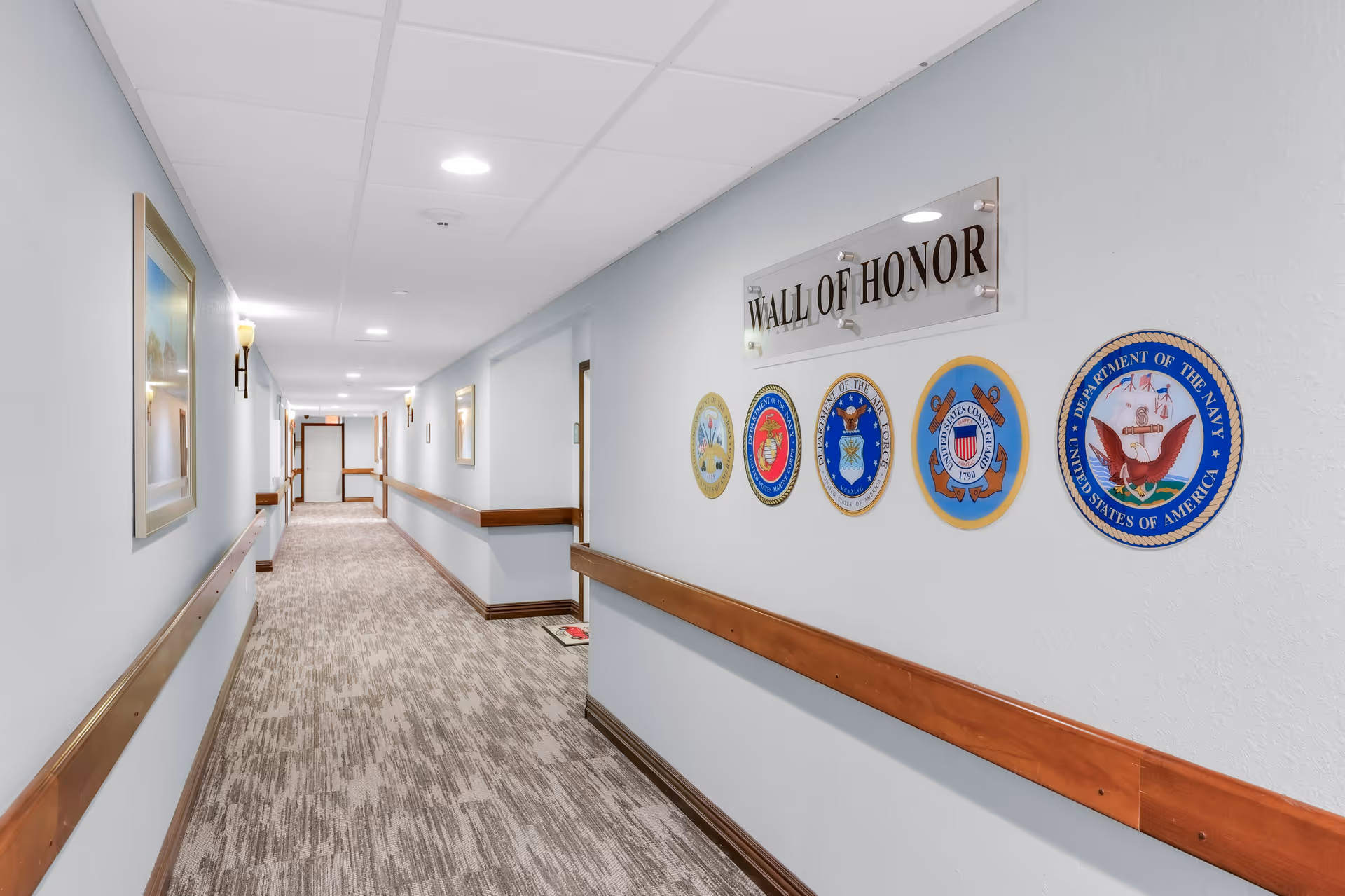 A well-lit interior hallway with handrails and a 'Wall of Honor' display of military seals on the right wall.