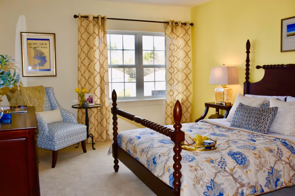 Bright furnished bedroom with a wooden four-poster bed, patterned bedding, an armchair by a window, and a bedside table with lamp.