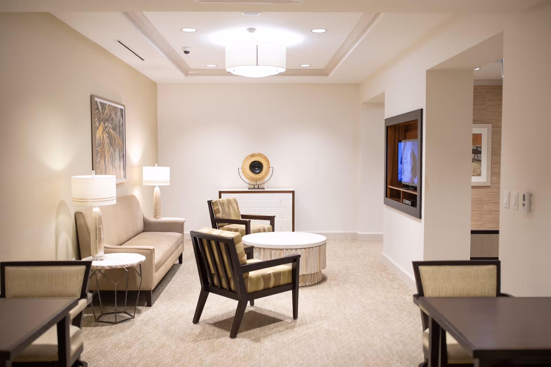 A well-lit living room area with a beige sofa, two armchairs with striped cushions, a round white coffee table, two side tables with lamps, a wall-mounted TV, and a decorative piece on a cabinet against the back wall. The room has neutral-colored walls and carpeted flooring.