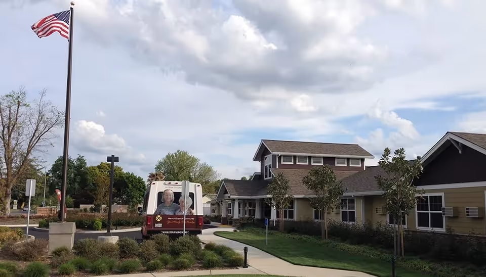 Exterior view of Amber Grove Place Memory Care facility with a clear sky and some clouds. The building has a combination of beige and brown siding with multiple windows and a sloped roof. A flagpole with an American flag is visible on the left side near a driveway where a shuttle bus with images of elderly people is parked. There are small trees and landscaped bushes along the sidewalk and driveway.
