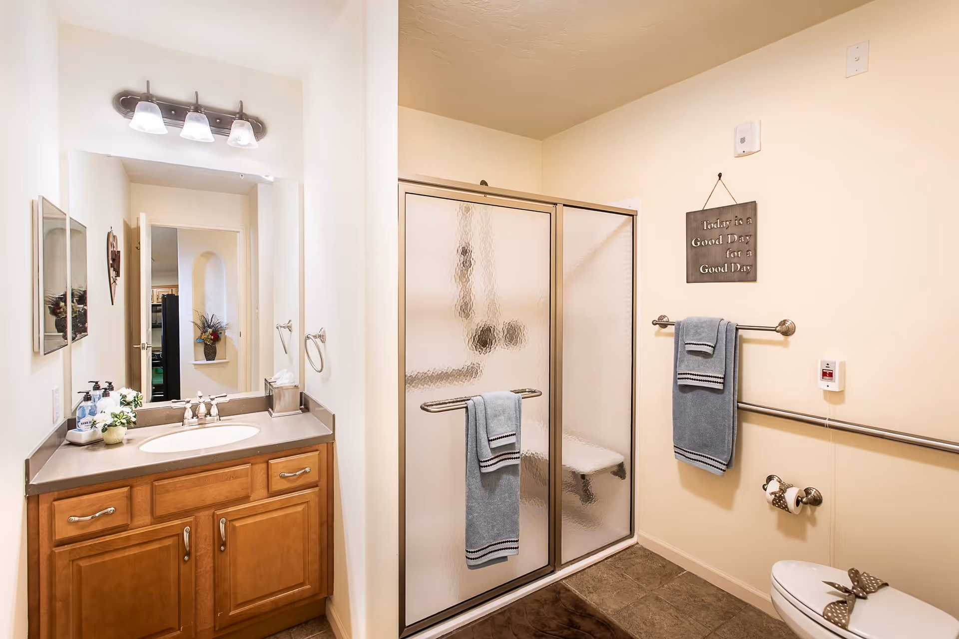 A clean and well-lit bathroom featuring a wooden vanity with a sink and mirror, a glass-enclosed shower with a built-in seat, and a toilet. Blue towels hang on towel bars, and a small decorative sign on the wall reads 'Today is a Good Day for a Good Day.' The bathroom has beige walls and tiled flooring.