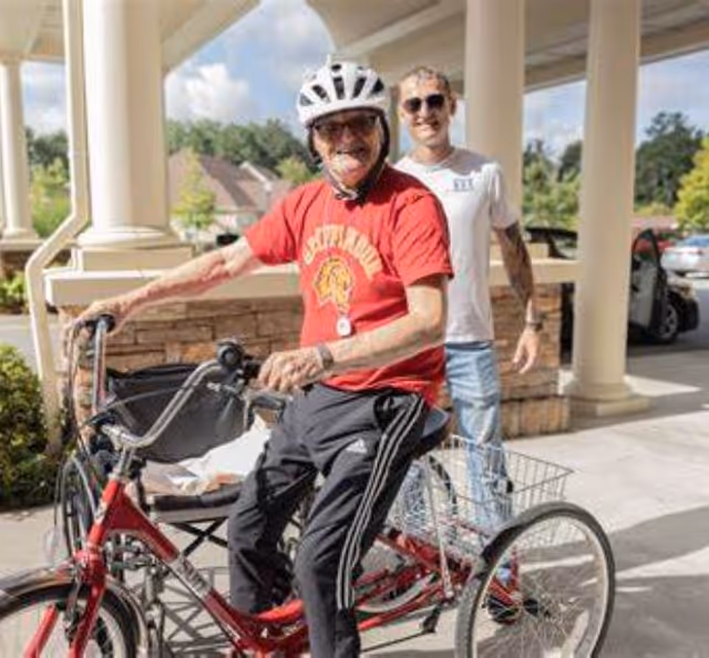 An elderly man wearing a white helmet and red shirt is sitting on a red tricycle outside a building with large columns. A younger man wearing sunglasses and a white t-shirt stands behind him, smiling. The scene is bright and sunny with greenery and parked cars in the background.