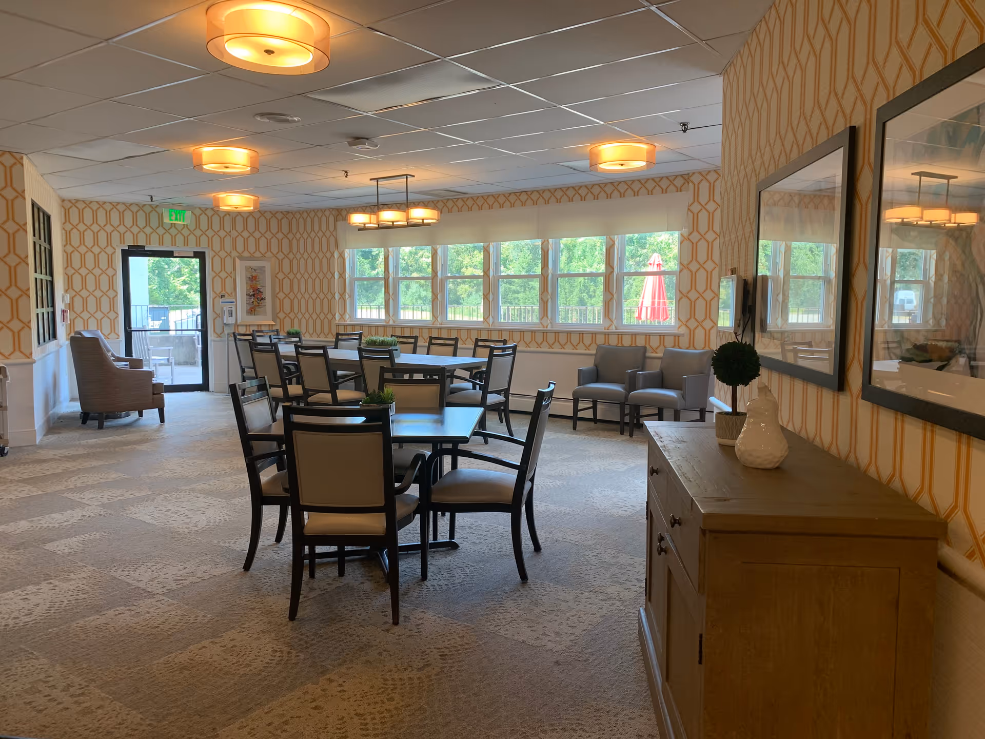 A spacious dining room with multiple tables and chairs arranged neatly. The room has patterned wallpaper, large windows letting in natural light, and modern ceiling lights. There are a few armchairs near the windows and a wooden cabinet with decorative items on the right side.