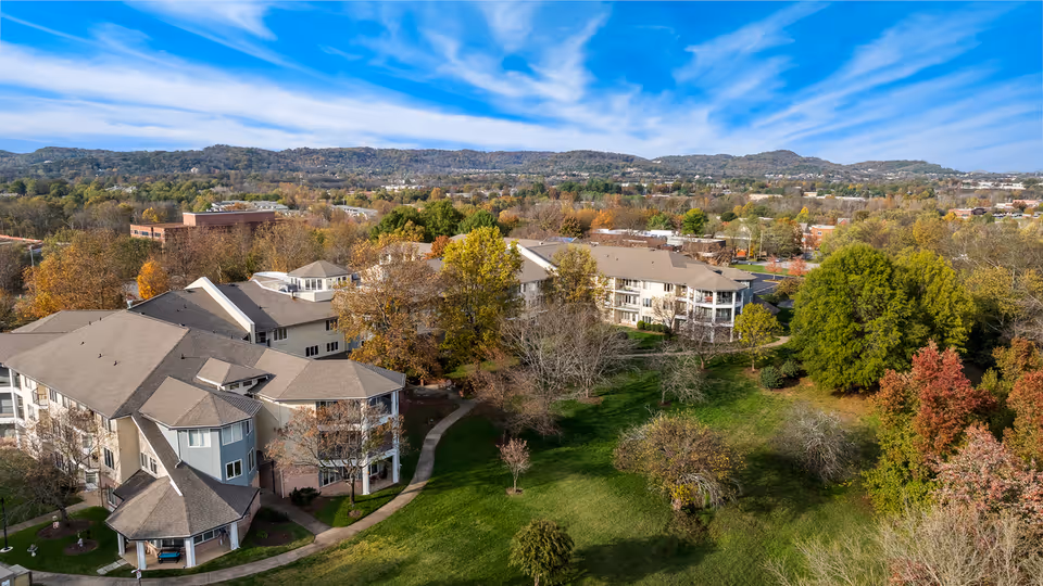 Aerial view of Manor at Steeplechase, a senior living community surrounded by green lawns, trees with autumn foliage, and walking paths. The buildings have beige and light blue exteriors with multiple windows and pitched roofs. Hills and a partly cloudy blue sky are visible in the background.