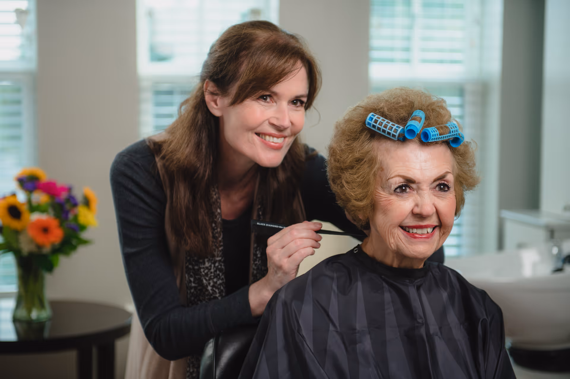 A smiling elderly woman with blue hair rollers in her hair sits in a salon chair wearing a black cape, while a hairstylist with long brown hair stands behind her, smiling and holding a comb. In the background, there is a vase with colorful flowers on a table and a window with blinds.