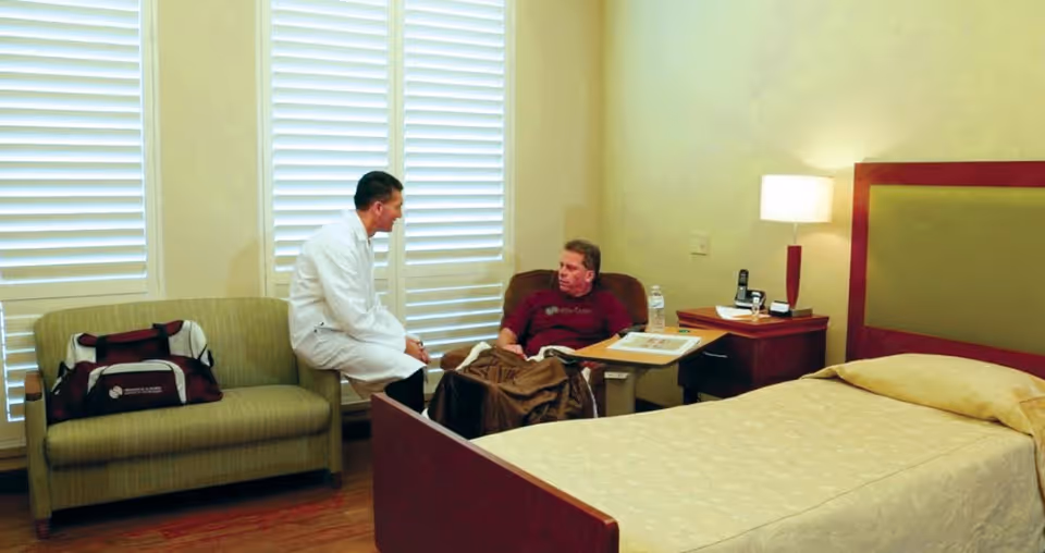 A resident seated in a recliner talks with a person in a white coat in a private bedroom-style care room with a bed, nightstand, lamp, and sofa.