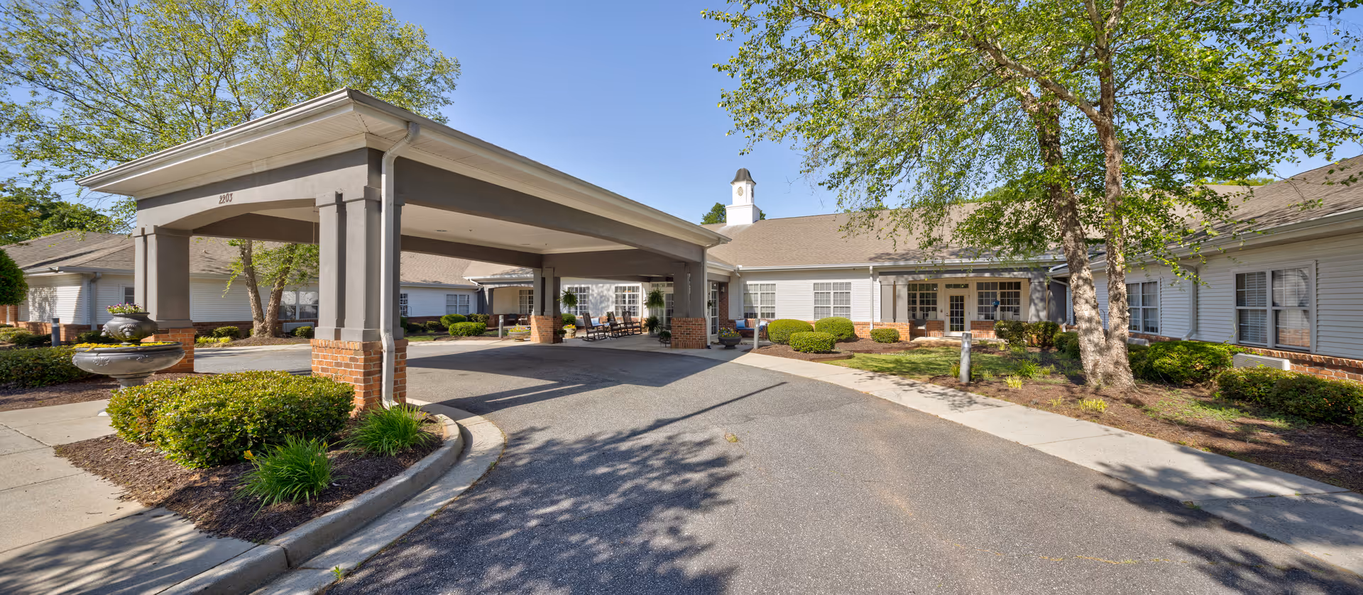 Front exterior view of TerraBella Marchbanks facility showing a covered driveway entrance with brick and beige pillars, surrounded by landscaped bushes, trees, and a clear blue sky.