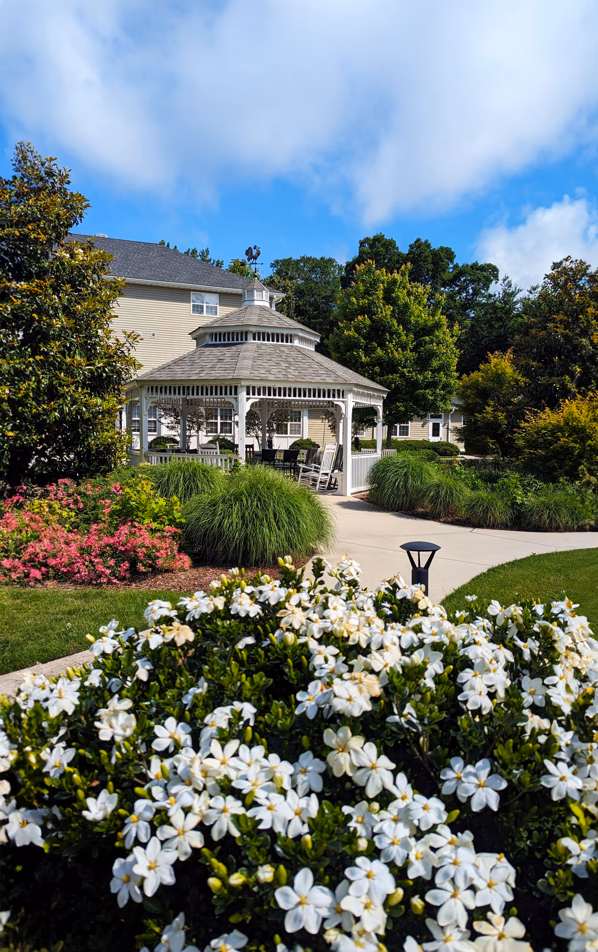 A bright outdoor garden area at Mennowood Retirement Community featuring a white gazebo with seating, surrounded by lush green bushes, blooming white and pink flowers, and a paved walkway. The background shows part of a beige building and tall trees under a partly cloudy blue sky.