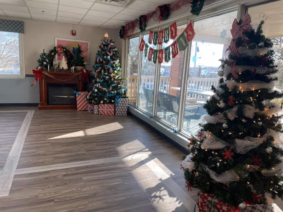 Sunlit common room decorated for Christmas with two decorated trees, a fireplace, wrapped presents, and large windows displaying a 'Merry Christmas' banner.