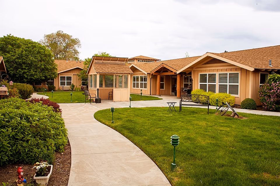 Outdoor view of Wildflower Lodge Senior Living showing a well-maintained garden with green grass, bushes, and a paved walkway leading to a tan-colored building with multiple windows and a small covered seating area.
