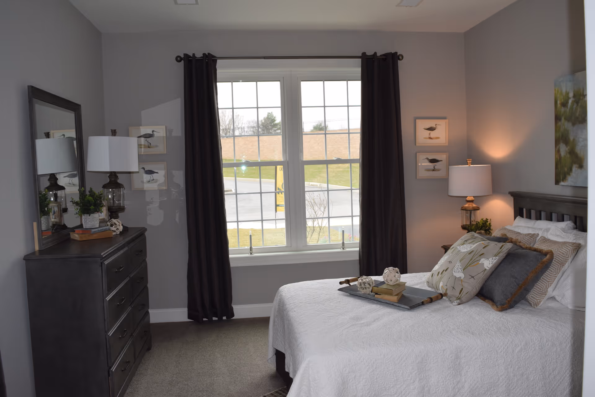 A cozy bedroom with a large window covered by dark curtains. The bed is made with white bedding and decorated with multiple pillows and a tray holding decorative items and books. To the left, there is a dark wooden dresser with a lamp, a small plant, and a mirror above it. On the right side of the bed, there is a nightstand with a lamp and a small plant. The walls are painted light gray and decorated with framed bird artwork.