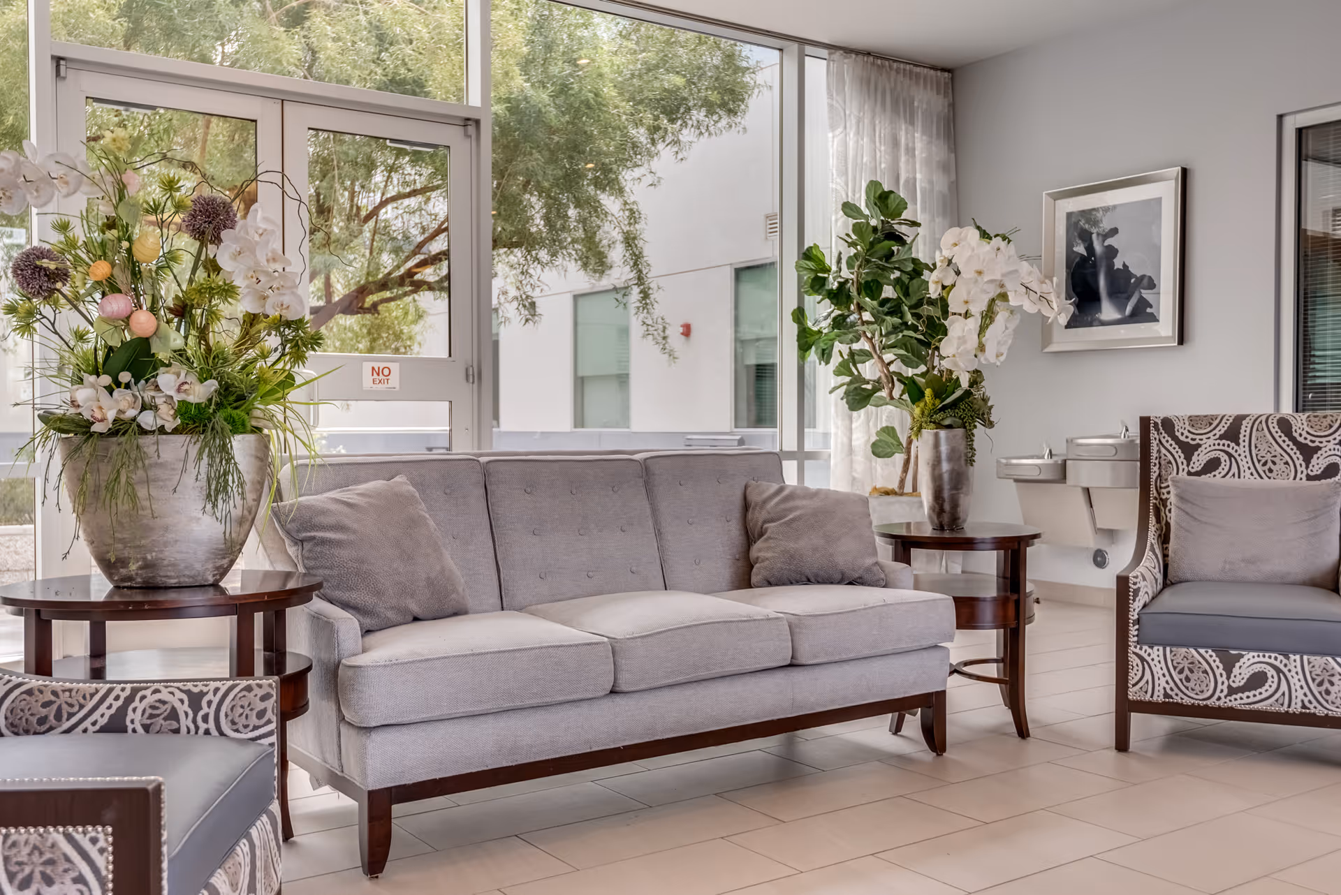 Bright seating lounge with a gray sofa, patterned armchairs, large floral arrangements, and floor-to-ceiling windows overlooking trees.
