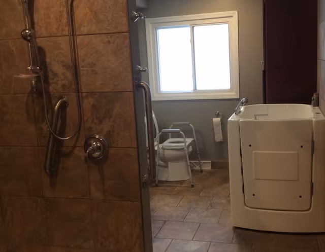A senior-accessible bathroom with a tiled walk-in shower with grab bars, a walk-in bathtub, and a raised toilet with a commode chair under a window.