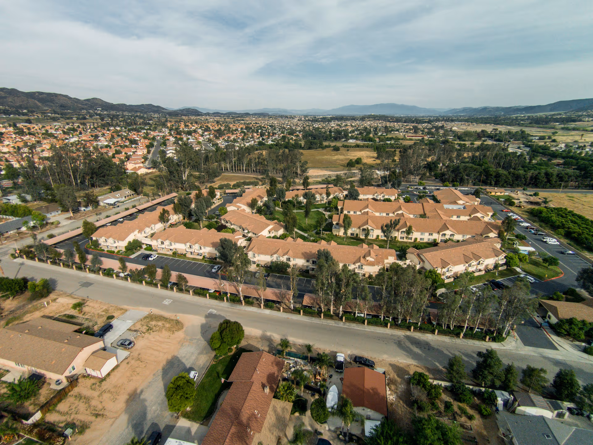 Aerial view of a senior assisted living facility with multiple buildings featuring red-tiled roofs, surrounded by trees and parking areas. The facility is situated in a suburban area with mountains in the background under a partly cloudy sky.
