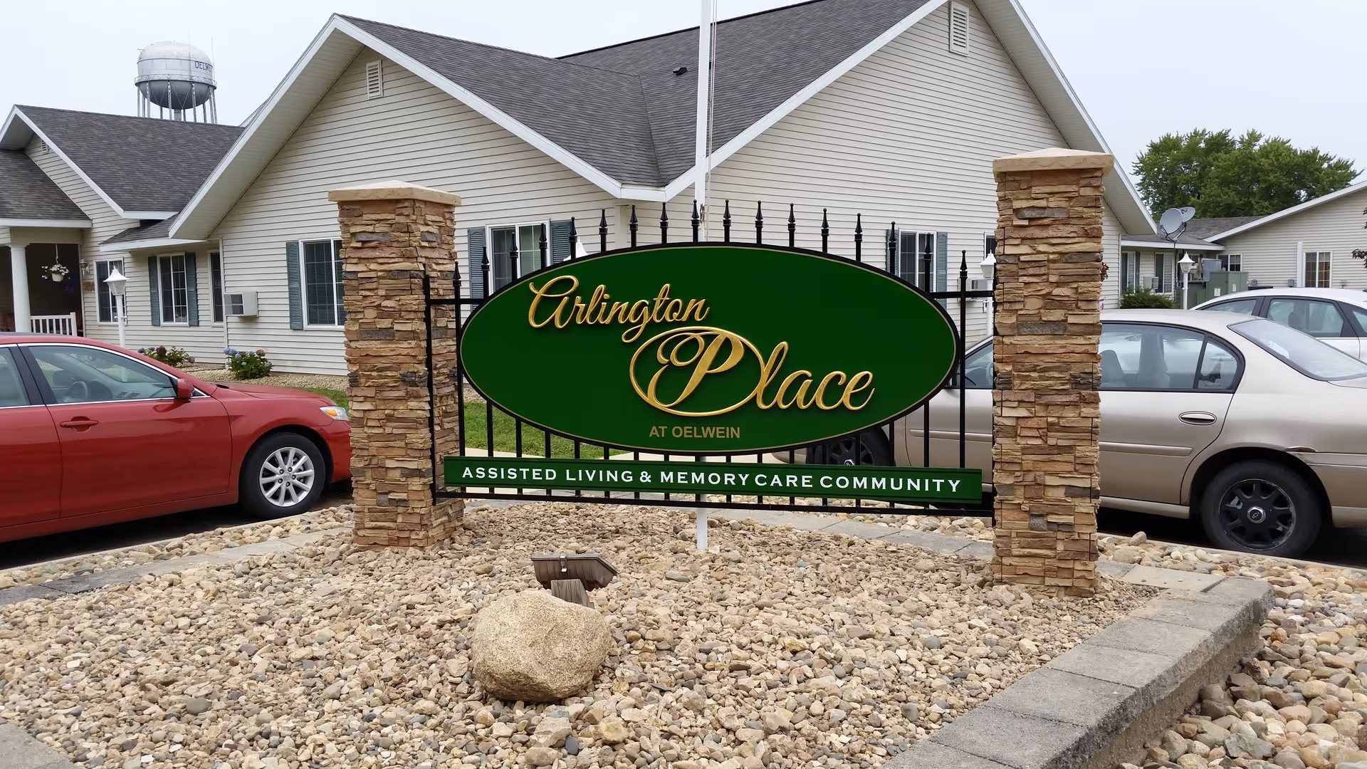 Outdoor view of Arlington Place at Oelwein assisted living and memory care community sign mounted on a black iron fence between two stone pillars, with a gravel bed in front and parked cars and residential buildings in the background.