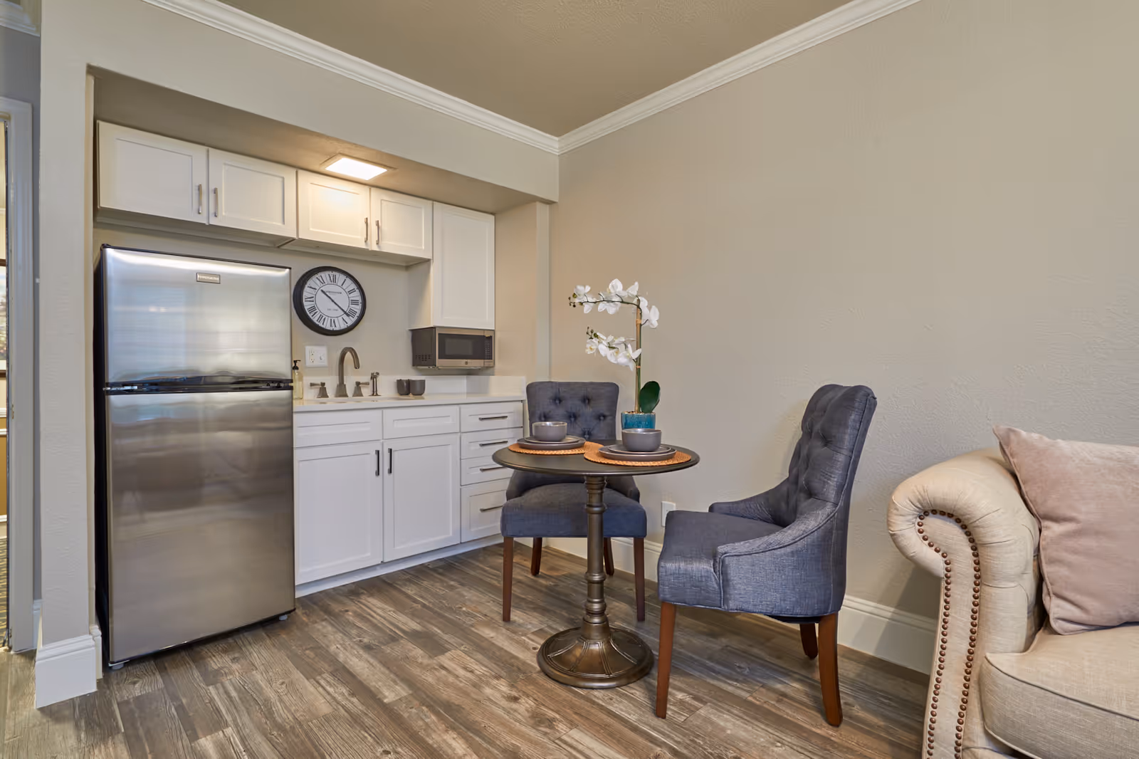 A small kitchenette area with white cabinets, a stainless steel refrigerator, a wall-mounted microwave, and a round table set for two with gray upholstered chairs and a white orchid centerpiece. Part of a beige sofa with a pink pillow is visible on the right.