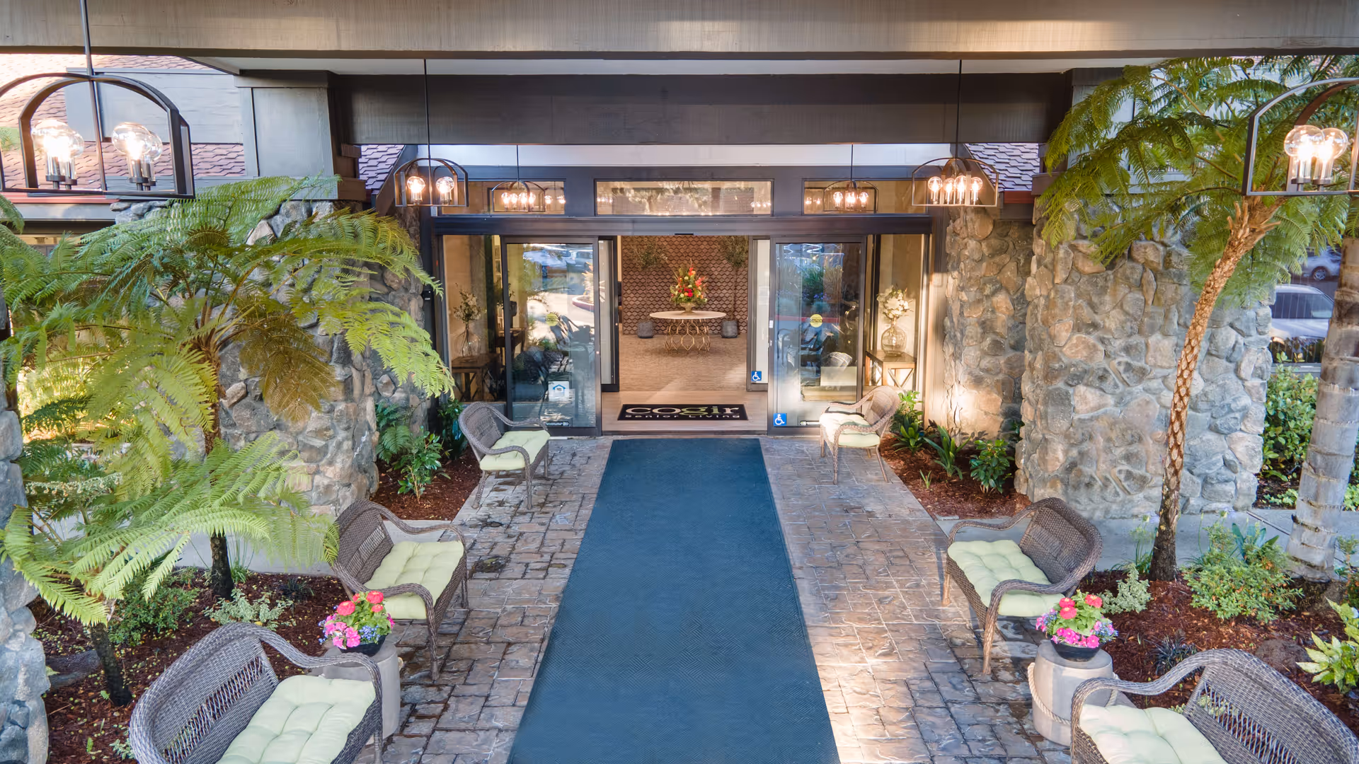 Entrance area of Cogir of Fremont senior living facility featuring a covered walkway with stone pillars, cushioned wicker chairs, potted plants, and hanging lantern-style lights leading to glass automatic doors with a welcome mat inside.