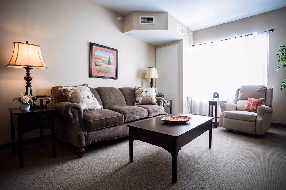 Well-lit living room with a sofa, armchair, coffee table and side tables with lamps by a large window.