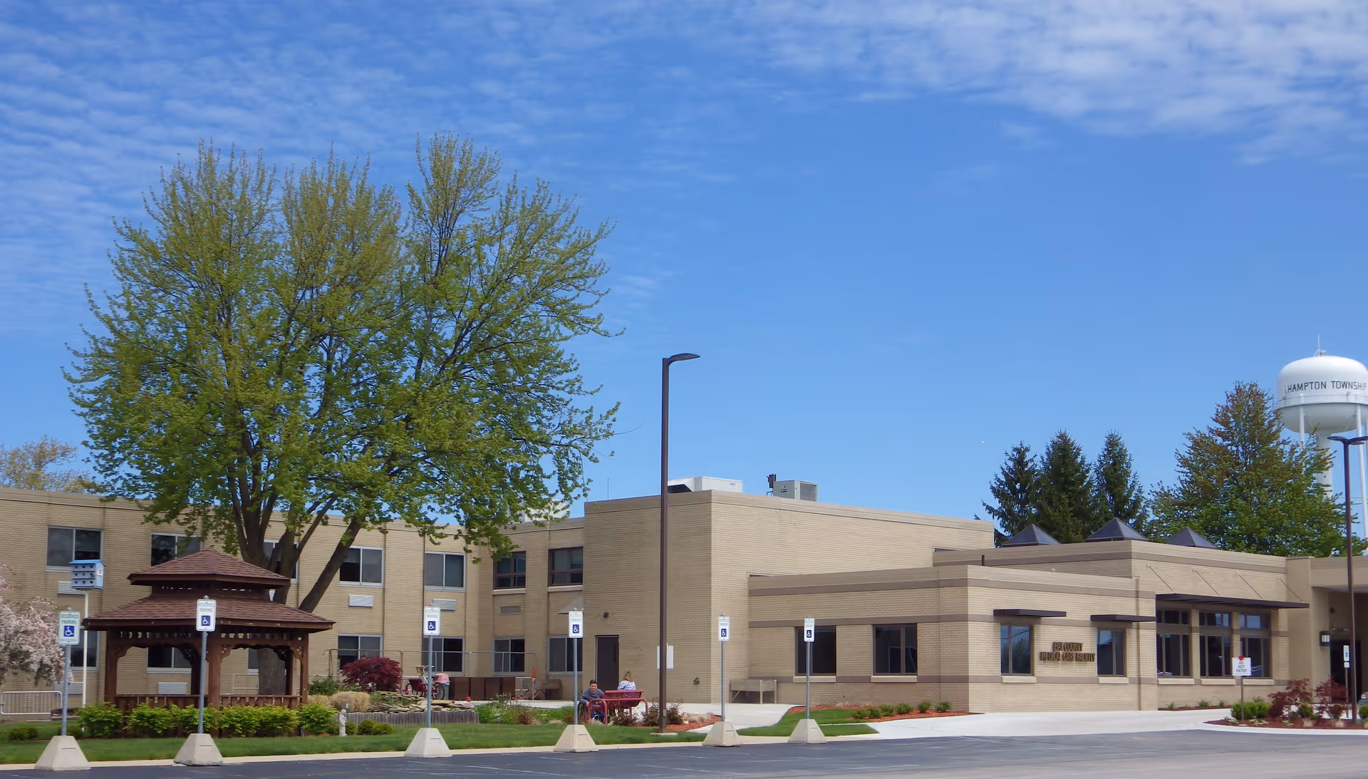 Front exterior of a low-rise medical care facility with a gazebo, large tree, and a water tower under a blue sky.