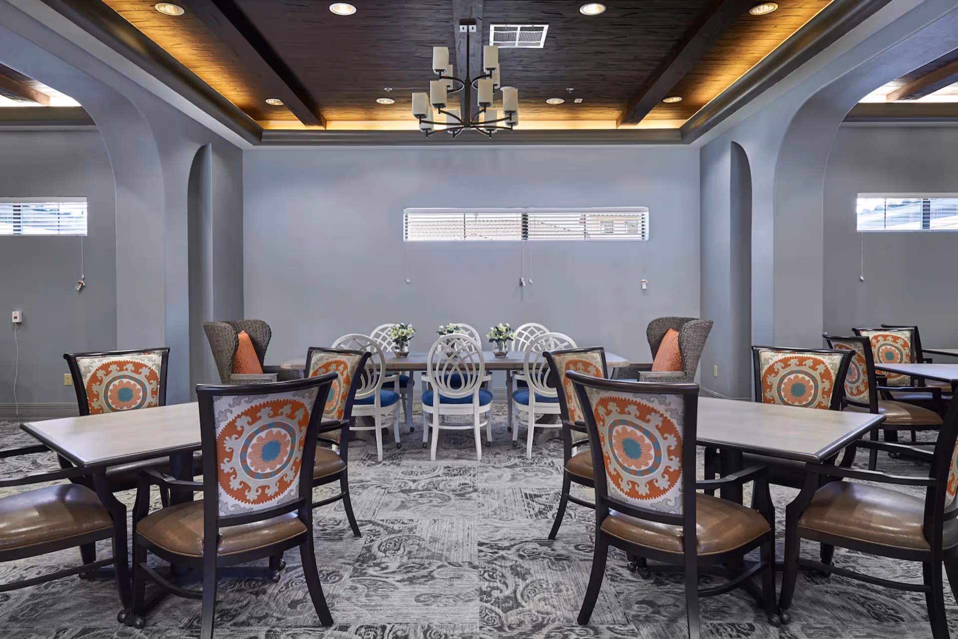 A dining room with multiple tables and chairs. The chairs have patterned backs with orange, blue, and beige designs. There are two wicker armchairs with orange cushions near the back wall. The ceiling features wooden beams and a modern chandelier with multiple lights. The walls are painted gray, and there is a narrow horizontal window with blinds on the back wall. The floor has a patterned carpet in shades of gray.