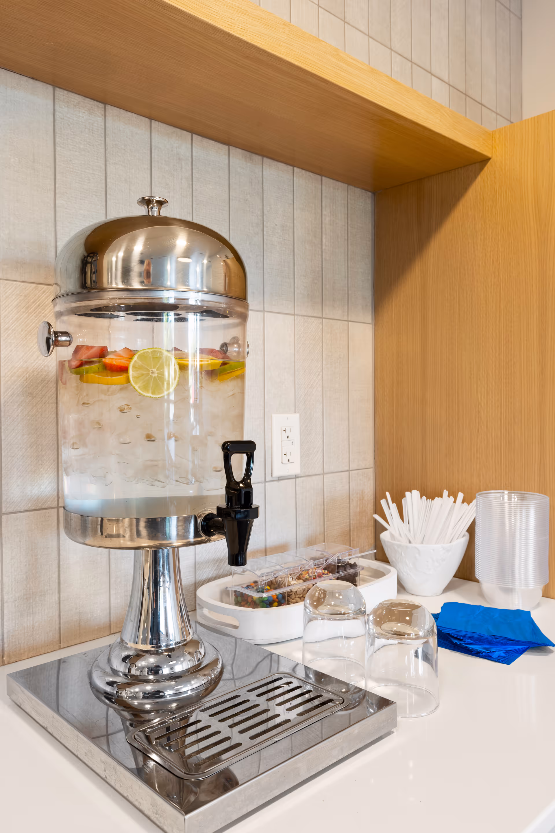 A countertop beverage dispenser filled with ice and fruit slices next to cups, straws, and napkins.