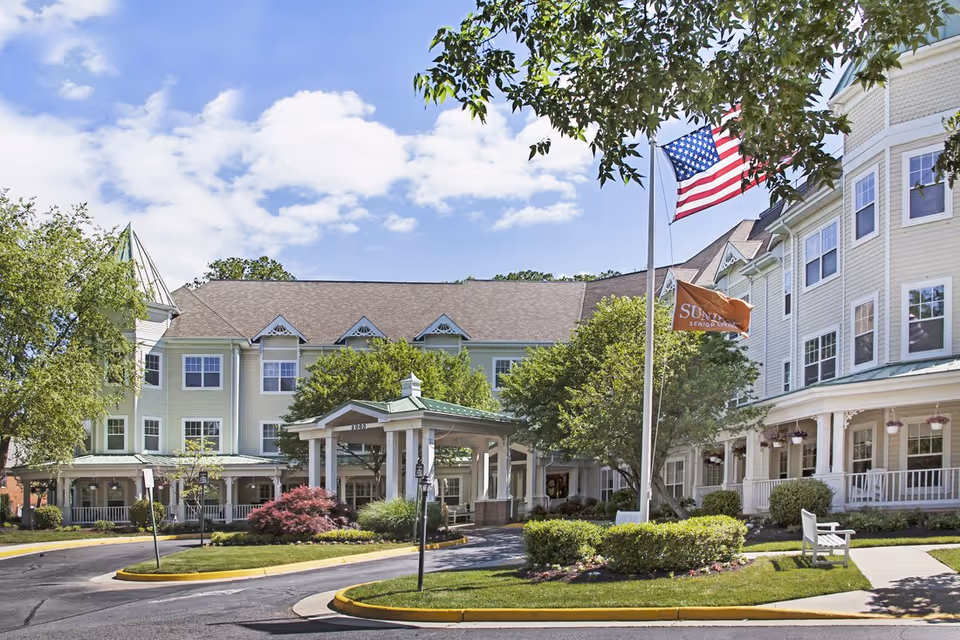 Front exterior of Sunrise at Hunter Mill senior living building with a covered entrance, American flag, landscaping, and circular driveway.