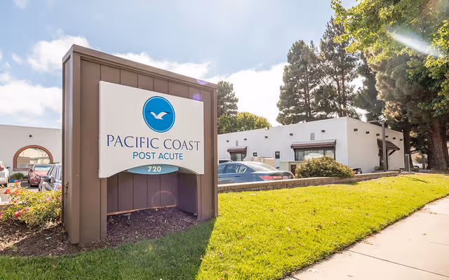 Outdoor view of the Pacific Coast Post Acute facility sign with a white building, parked cars, trees, and a sidewalk under a partly cloudy sky.