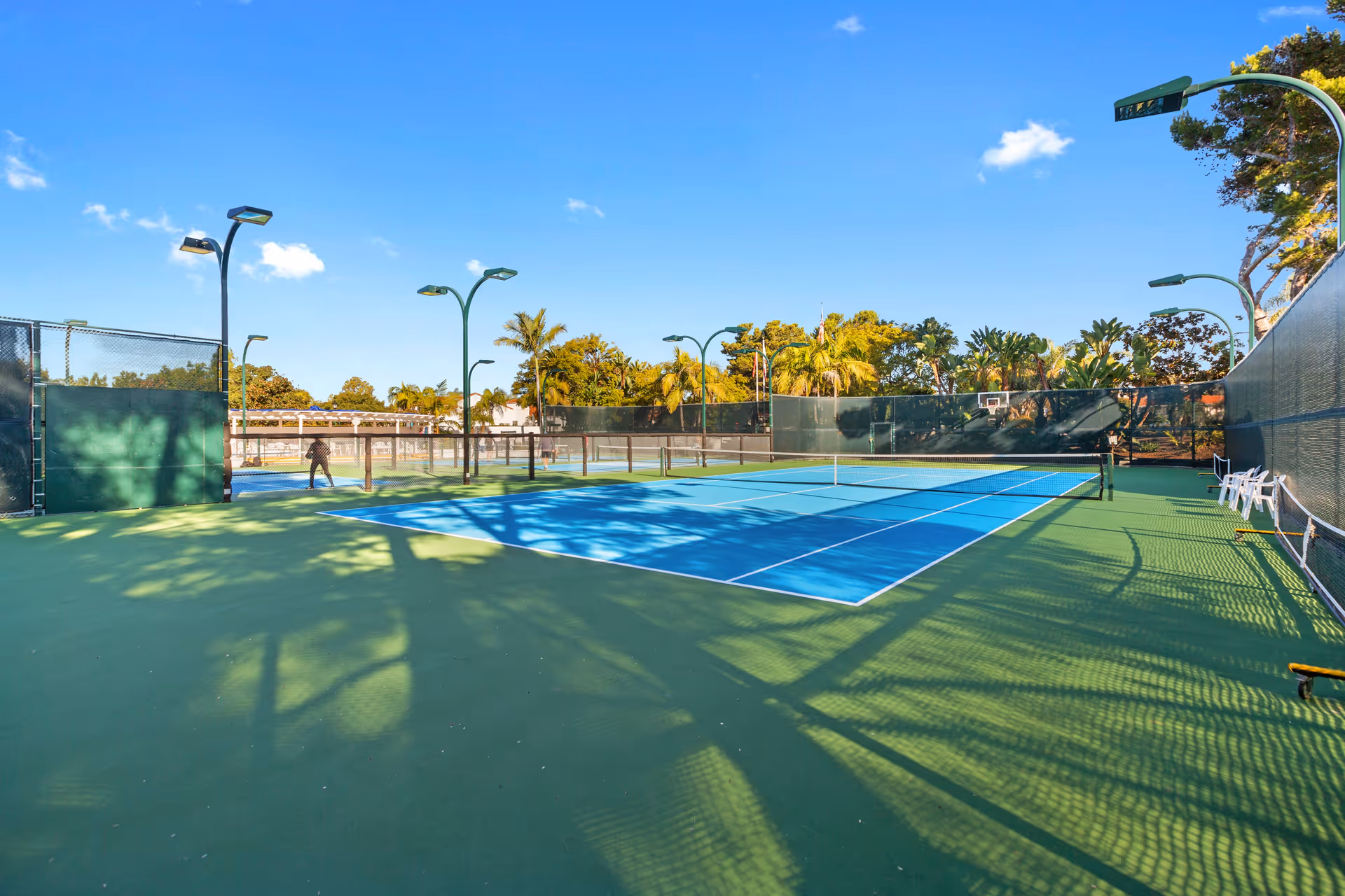 Outdoor tennis courts with blue and green surfaces surrounded by tall green fences and multiple light poles. Palm trees and other greenery are visible in the background under a clear blue sky.
