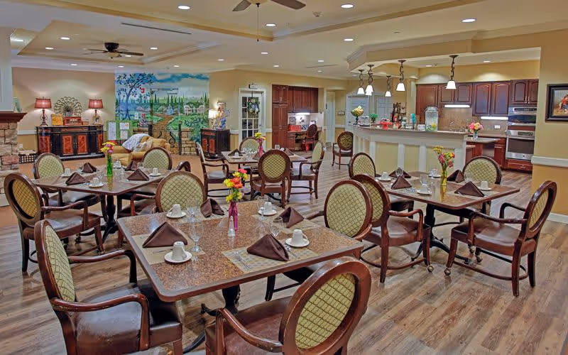 A spacious dining room with multiple tables set with cups, glasses, and brown napkins. Each table has a small vase with colorful flowers. The room features wooden chairs with cushioned seats and backs, a ceiling fan, and warm lighting. In the background, there is a kitchen area with wooden cabinets, a countertop, and appliances. A mural and decorative furniture are also visible along the back wall.