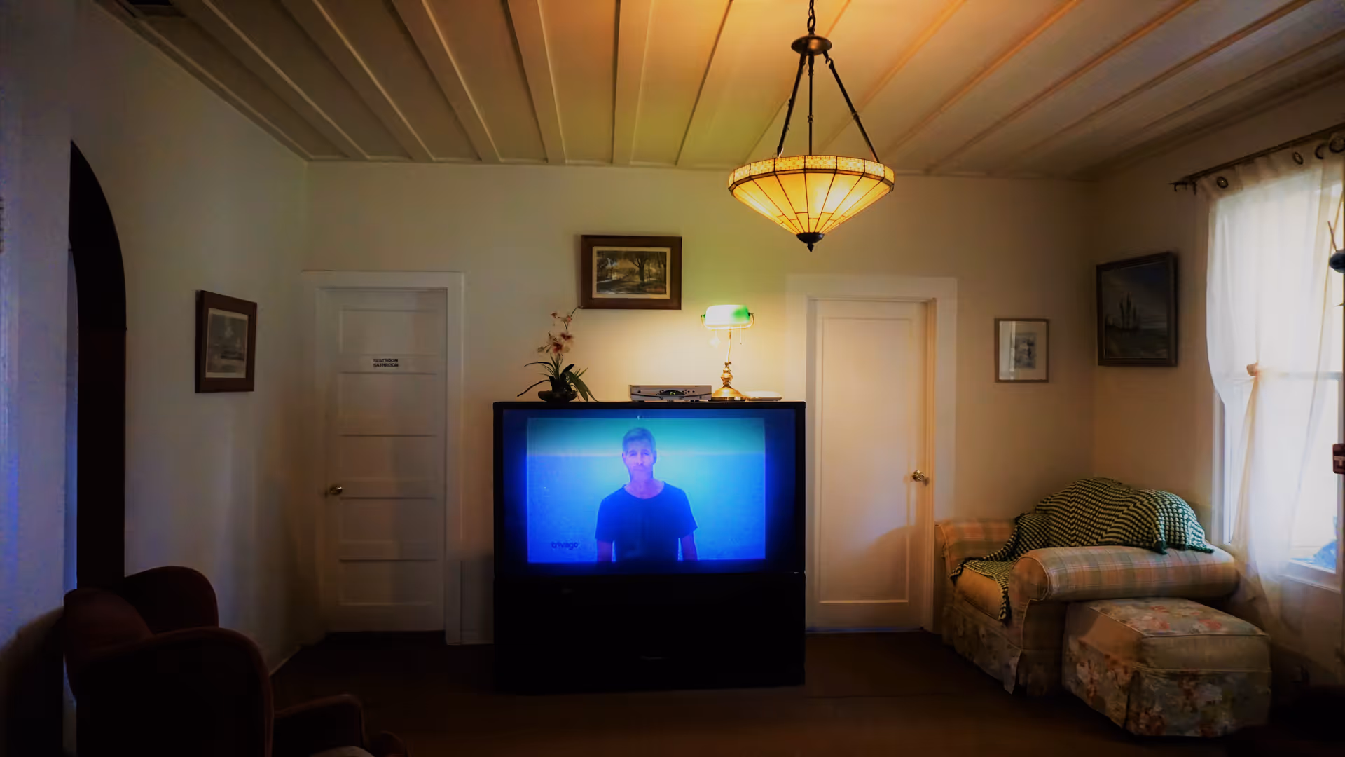 A cozy living room with a large older television showing a person, upholstered chairs and sofa, wall art, and a hanging ceiling lamp.
