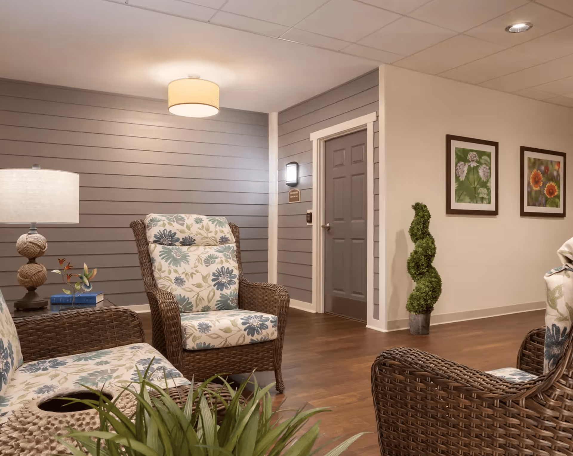 A cozy sitting area with wicker chairs featuring floral cushions, a table with a lamp and decorative items, a gray door on a wall with horizontal paneling, two framed floral pictures on the adjacent wall, and a spiral-shaped topiary plant in a pot on a wooden floor.
