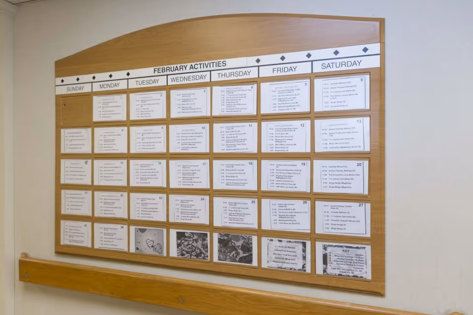 A wooden-framed bulletin board mounted on a wall displaying a calendar of February activities with daily schedules listed under each day of the week from Sunday to Saturday.