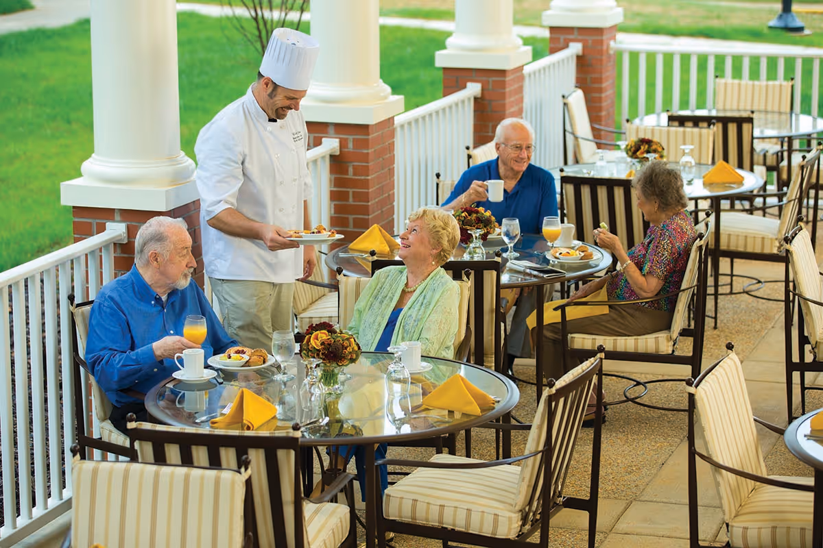 Seniors enjoying a meal on a covered outdoor patio while a chef serves them.