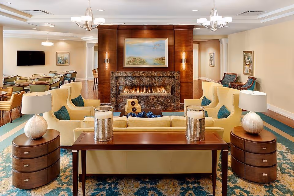 A cozy living room area in a senior living facility featuring a modern fireplace with a stone surround and a painting above it. The room has beige walls, wooden floors, and a patterned blue and beige rug. There are several yellow armchairs with teal cushions arranged around a wooden coffee table, flanked by two round wooden side tables with lamps. In the background, there is a dining area with tables and chairs, a wall-mounted TV, and additional seating along the walls with framed artwork.