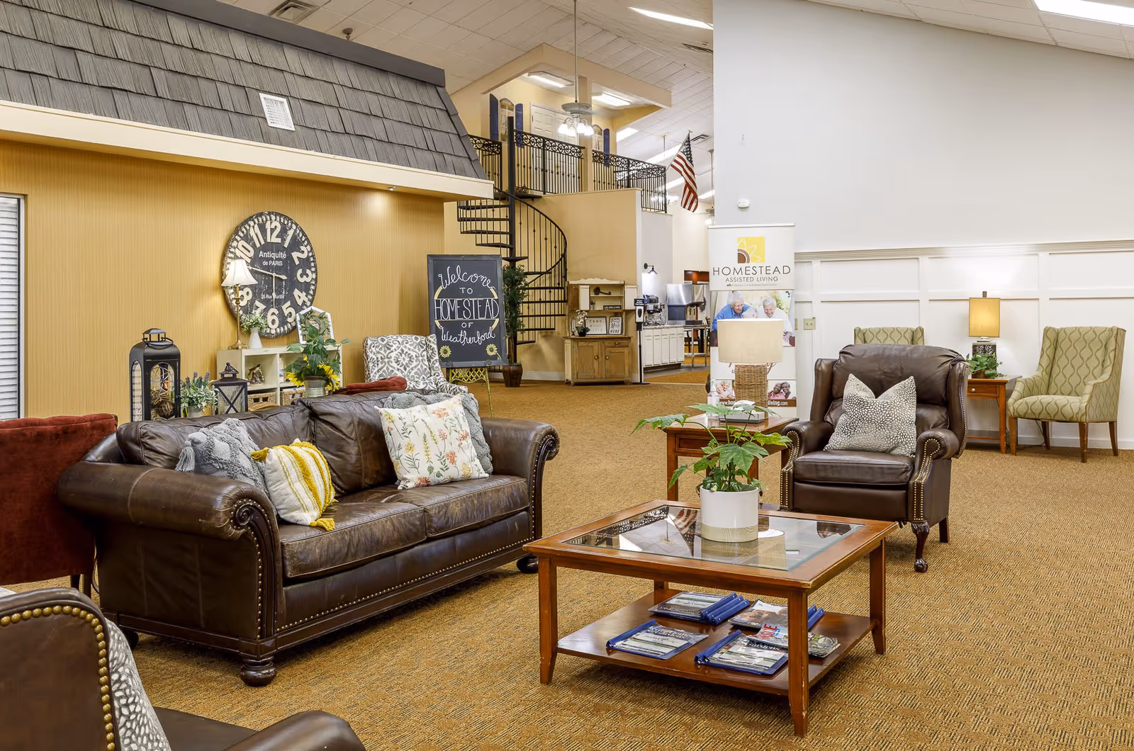 A cozy assisted living facility common area with a brown leather sofa and armchair, a wooden coffee table with magazines, decorative pillows, a potted plant, and a large wall clock. In the background, there is a spiral staircase, a welcome sign for Homestead of Weatherford, and a banner for Homestead Assisted Living. The room has warm lighting and carpeted floors.