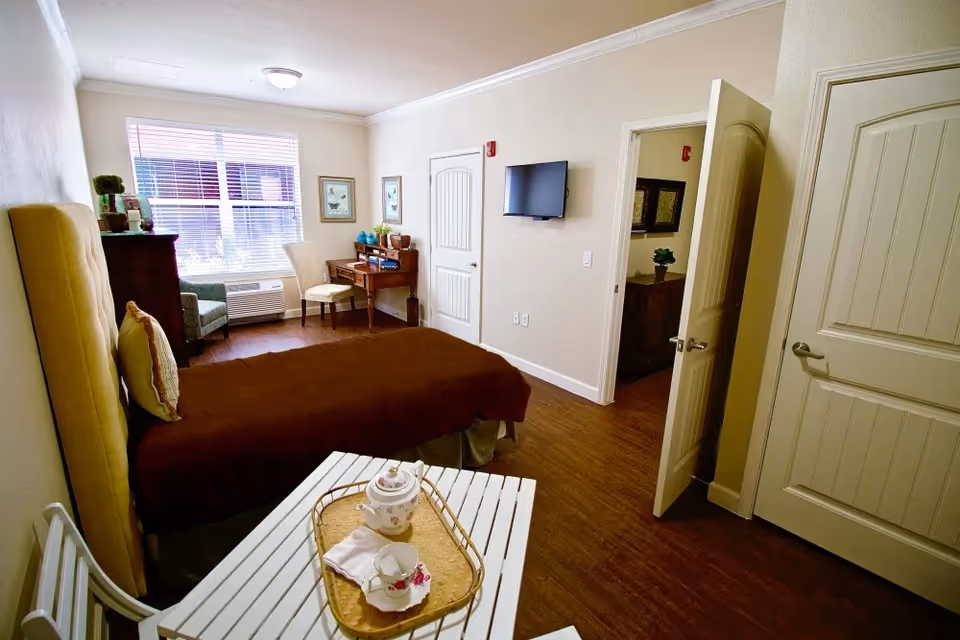 Bright senior living bedroom with a bed, small table and tea set, desk and chair by a window, wall-mounted TV, and an open door.