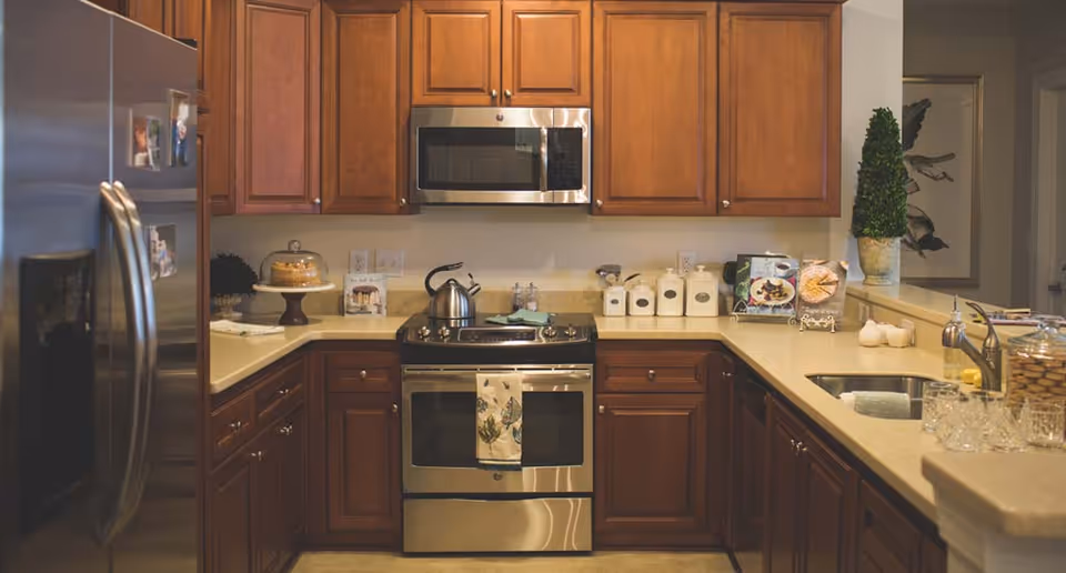 A modern kitchen with wooden cabinets, a stainless steel refrigerator, oven, and microwave. The countertops have various items including a kettle on the stove, a cake stand with a cake, canisters, a small potted plant, and some glassware near the sink.