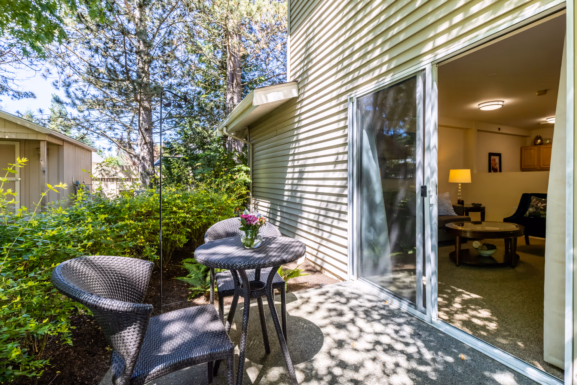 Small shaded patio with wicker table and chairs and a vase of flowers next to a sliding glass door opening into a living room.