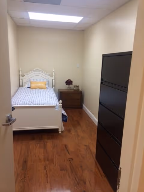 A small bedroom with a white wooden bed featuring a blue and white patterned bedspread and a yellow pillow. Next to the bed is a wooden nightstand with a vase of flowers and a small decorative item. The room has wooden flooring, beige walls, a black dresser on the right side, and a ceiling light panel.