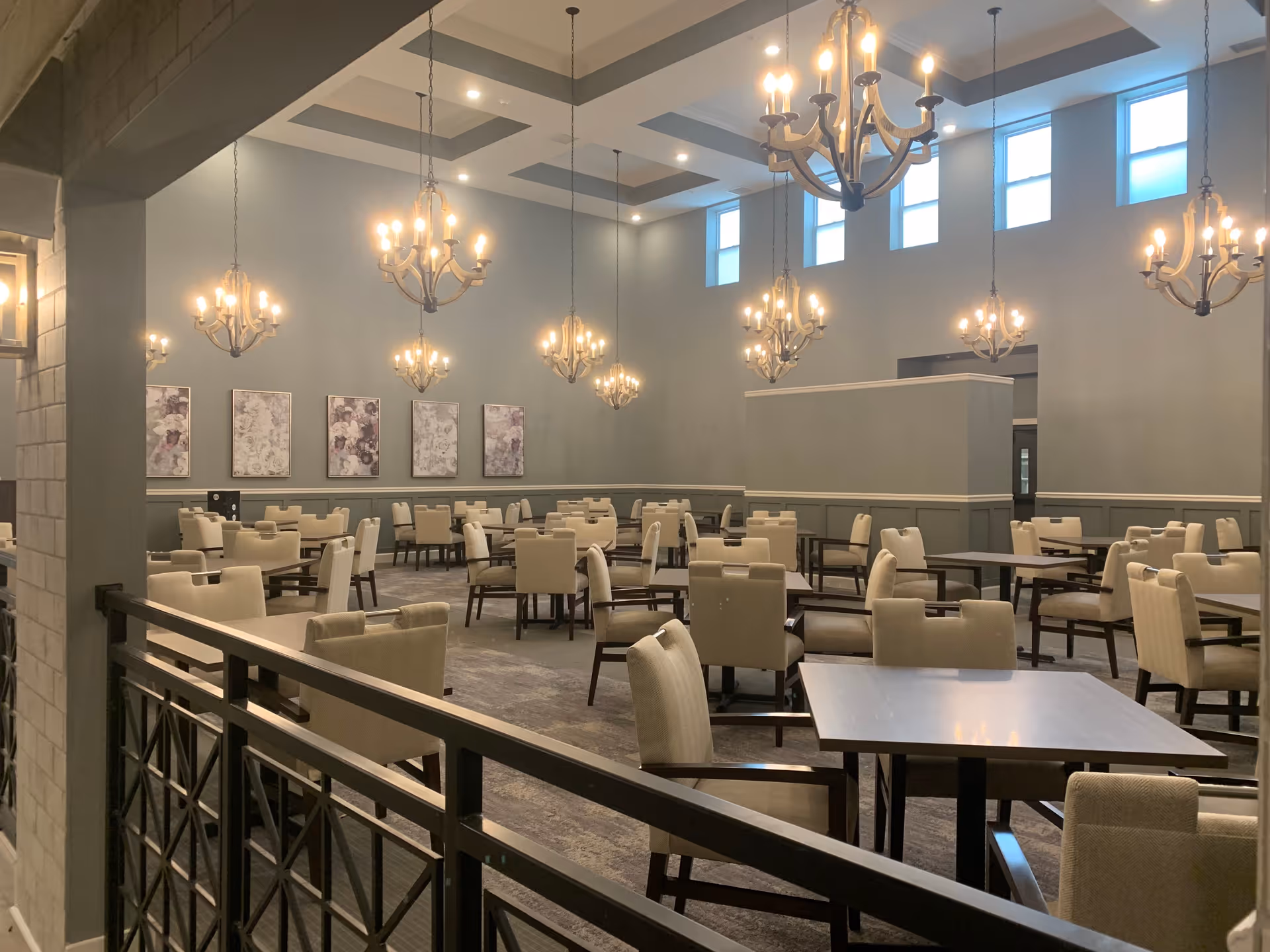 A spacious dining room with multiple square tables and beige upholstered chairs arranged neatly. The room features high ceilings with several elegant chandeliers hanging down, and tall windows near the ceiling letting in natural light. The walls are painted gray and decorated with framed artwork.