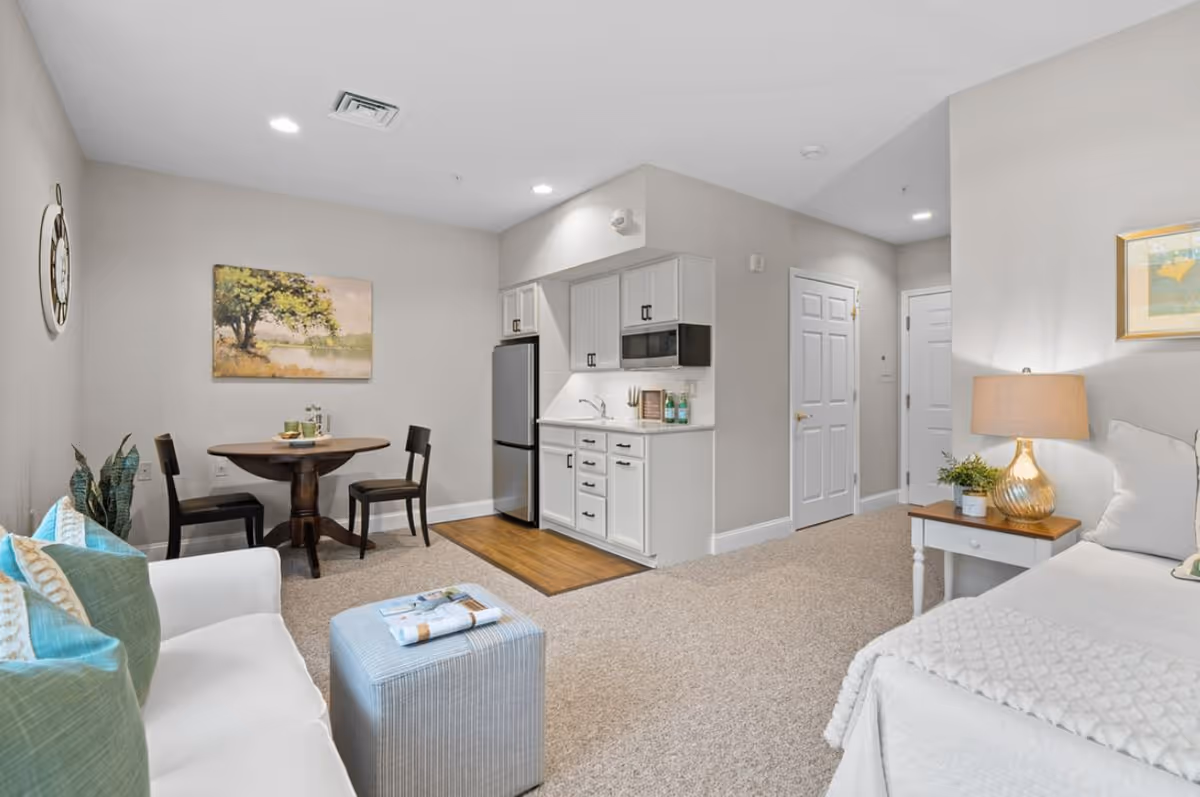 Bright studio apartment interior with a small kitchenette, round dining table, sofa, and bed in a furnished senior living unit.