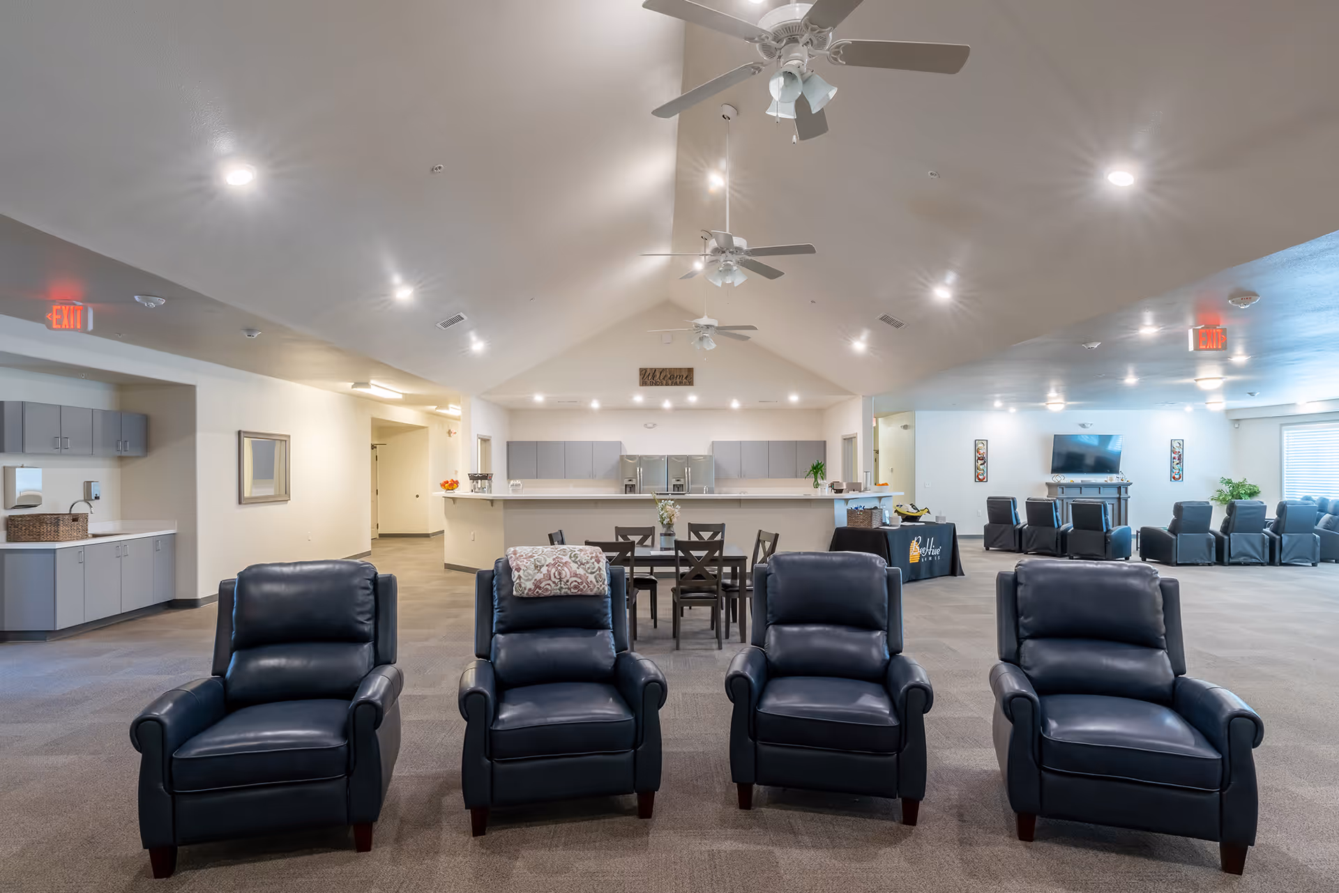 A spacious, well-lit common area in a senior living facility featuring four black leather armchairs in the foreground, a dining table with chairs in the middle, and a kitchen area with gray cabinets and stainless steel appliances in the background. Ceiling fans and recessed lighting illuminate the room, and additional seating is visible near a TV on the right side.